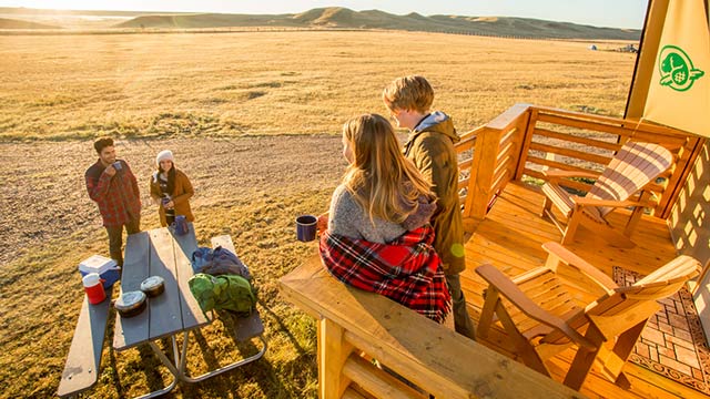Deux jeunes adultes sont sur la terrasse d'une tente oTENTik et discutent avec deux jeunes adultes qui sont près de la table de pique-nique au parc national des Prairies.