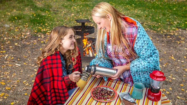 Une mère et sa fille emmitouflées dans des couvertures près d'un feu de camp se servent une boisson chaude au parc national de Prince Albert.