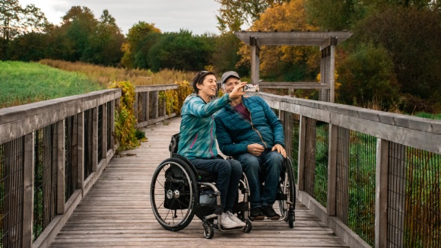 Deux adultes en fauteuil roulant se prennent en photo sur un trottoir de bois.